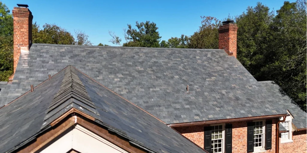 Slate roof with copper flashing on a historic brick home with two chimneys and mature trees in the background